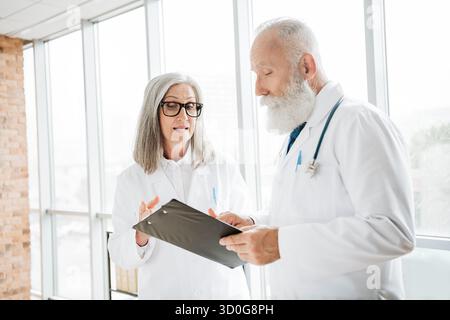 Two elderly doctors review a patient file and discuss treatment in a bright clinic for health care and professional collaboration Stock Photo