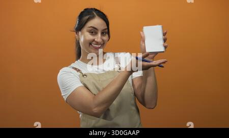 Woman in apron holding a notepad and pen, smiling and showing her hand in an orange studio; friendly service. Stock Photo