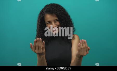 Young hispanic woman with open palms beckons in studio against teal wall with warm smile; friendly invitation. Stock Photo