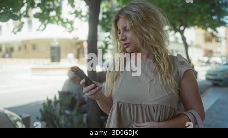Young blonde woman holding smartphone on street under tree, wearing beige sundress and handbag on elbow while glancing at passing cars; pensive waitin Stock Photo