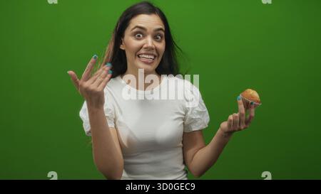 Young hispanic woman holding muffin beckons with open hand in green studio, smiling broadly and speaking to camera; playful invitation. Stock Photo
