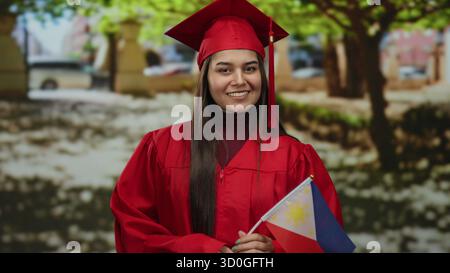 Woman in red graduation gown holding philippine flag in a park setting, symbolizing accomplishment, pride, and national identity. Stock Photo