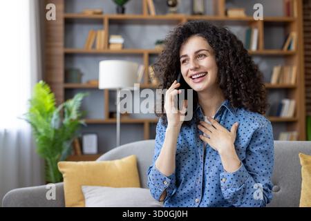 Young woman with curly hair smiling and talking on a mobile phone, relaxed on a cozy living room couch at home, sharing joyful conversation and feeling contentment Stock Photo