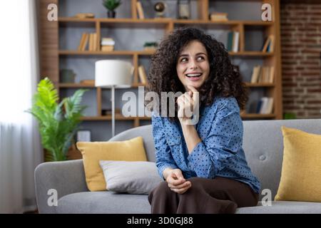 Happy young woman with curly hair sitting on a cozy sofa, smiling and daydreaming, looking away, reflecting a moment of inspiration or new idea in a home living room Stock Photo