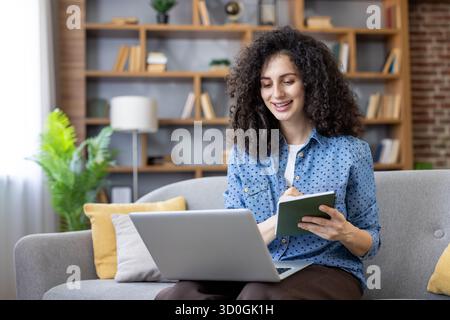 Young woman with curly hair confidently sitting on a sofa, taking notes in a notebook while using a laptop for remote work or online education in a cozy home environment Stock Photo