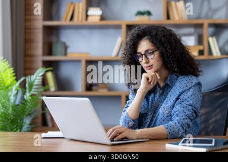 Young woman with curly hair and glasses concentrating while typing on a laptop at a wooden desk in a modern home office, symbolizing remote work and productivity Stock Photo