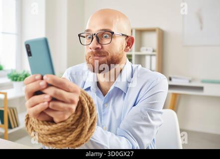 Addicted male employee with tied hands holding mobile phone sitting at the desk in office Stock Photo