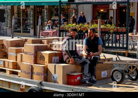 Two men in conversation sitting on cardboard boxes on a goods barge making deliveries via a canal in Venice,Italy Stock Photo