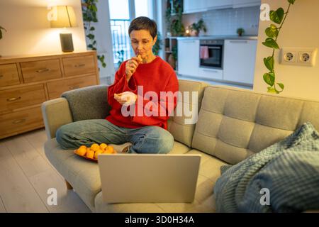 Relaxed mature woman watching movie. Content female eating mandarin laptop sofa, Downtime with snack Stock Photo