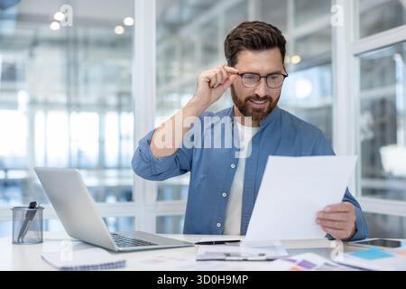 Young businessman adjusting spectacles while reading an important legal document or business report, analyzing information and working in a modern office environment Stock Photo