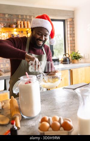 African American man wearing Santa hat whisking batter with whisk in glass bowl in kitchen Stock Photo