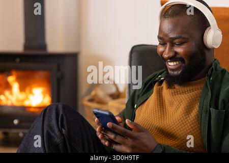 African American man sitting at home by wood stove with headphones, holding smartphone, copy space Stock Photo