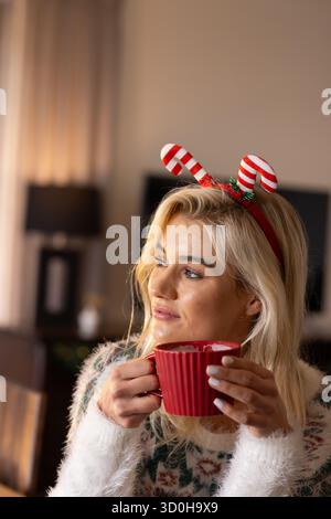 Woman wearing sweater, candy cane headband holding red mug near lips at table in living room Stock Photo