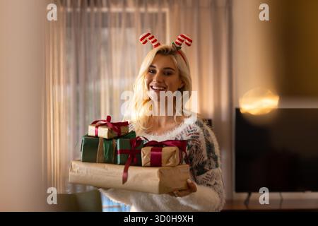 Smiling woman wearing holiday sweater, antler headband holding ribboned gift boxes in living room Stock Photo