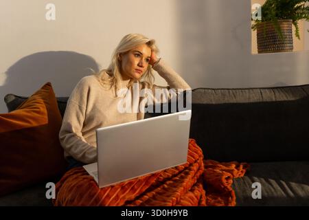 Woman sitting on dark gray sofa at home, holding silver laptop, noticing potted fern Stock Photo