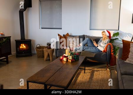 Woman reclining on sofa wearing Santa hat using tablet with gift boxes at home, copy space Stock Photo
