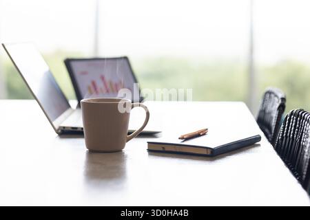 Steaming ceramic coffee mug on table beside open laptop, tablet showing chart, notebook with pen Stock Photo