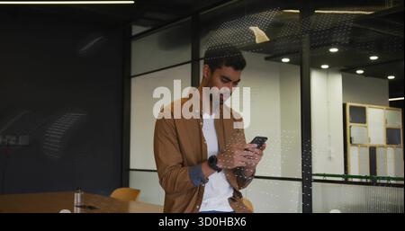 Leaning man in brown shirt checking smartphone at conference table in office with glass partitions Stock Photo