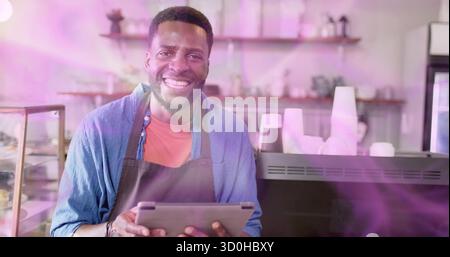 Smiling male barista wearing red shirt and dark apron, holding tablet at cafe, with coffee machine Stock Photo