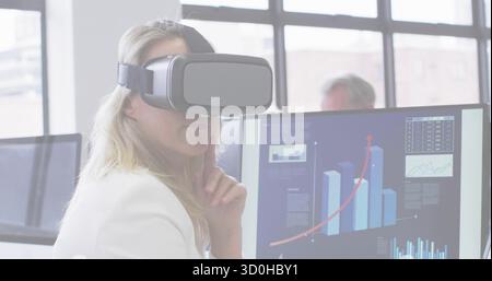 Resting female wearing VR headset, white blazer at office desk, with monitors showing upward trend Stock Photo