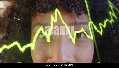 Showing close-up woman with nose ring gazing at fluctuating green line chart on glass in studio Stock Photo