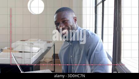 Smiling man analyzing documents and typing on laptop in bright office with pen and graph overlay Stock Photo