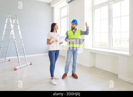 Builder in safety helmet talking to young woman in new house discussing renovation plans and repairs Stock Photo