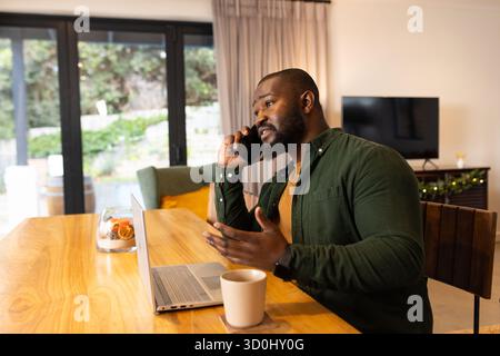 African American man talking on smartphone, using laptop at home dining table with mug, copy space Stock Photo