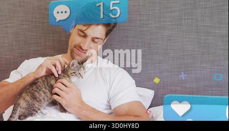 Reclining man in white t-shirt petting tabby kitten on bed in bedroom, with notification icons Stock Photo