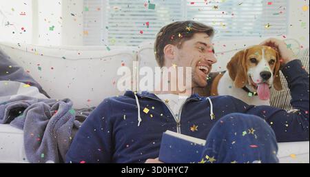 Reclining man wearing hoodie petting beagle on sofa in living room with blue book, confetti Stock Photo