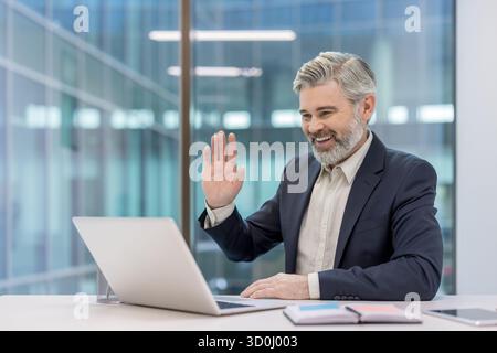 Smiling mature businessman having an online video conference call on a laptop, raising his hand to wave from a bright modern office with large glass windows Stock Photo