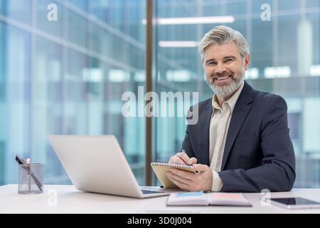 Mature businessman with gray hair and beard sitting at his office desk, confidently smiling at the camera while writing notes in a notebook next to a laptop Stock Photo