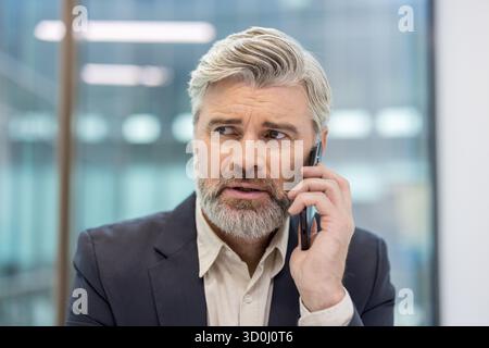 Mature executive man having a serious and concerned phone call, discussing a problem or critical information while looking away in an impersonal office setting Stock Photo