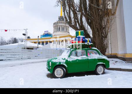 Small green retro car covered in snow with colorful wrapped presents on the roof, set against the backdrop of the main Kyiv VDNKh pavilion Stock Photo