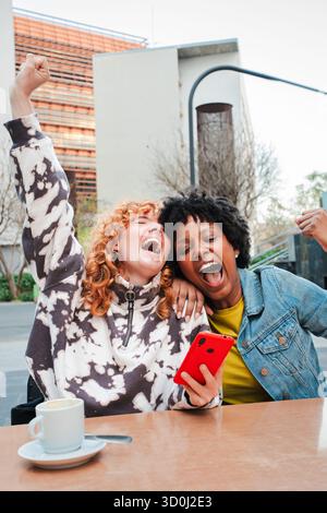 Two joyful friends celebrating a thrilling win together while excitedly using their smartphones and sharing their happiness in a lively atmosphere of Stock Photo