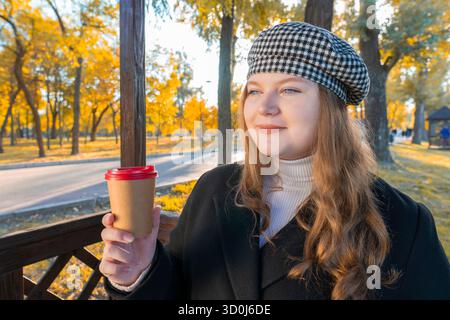 Close-up portrait of a smiling young woman in a checkered beret holding a paper coffee cup in an autumn park with golden foliage Stock Photo