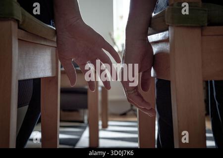 Two people sitting side by side reaching for each other’s hands under chairs indoors Stock Photo