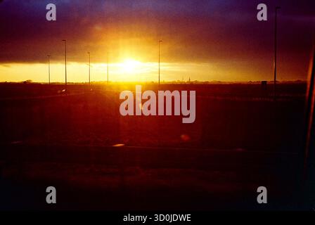 Analog contrasty shot of the setting sun breaking through clouds, captured from a moving car, highlighting dramatic light and motion. Stock Photo