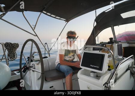 Man wearing a FPV drone goggles in the cockpit of a sailing yacht on calm open sea, surrounded by navigation equipment, modern boating and technology Stock Photo