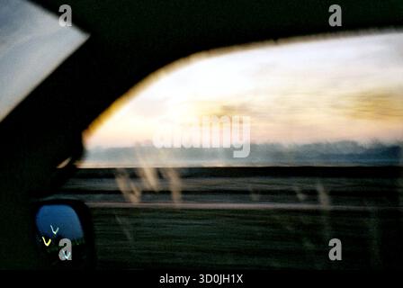 View through a moving van window at early morning dawn, river visible beyond the bridge railing, captured in an analog atmospheric style. Stock Photo