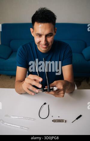 Vertical shot of cheerful man assembling wi-fi module by connecting antennas sitting at white table in home office, focused on ensuring optimal Stock Photo