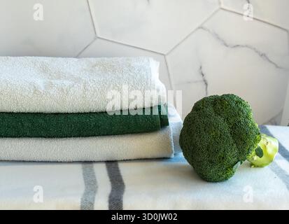 Fresh green broccoli vegetable on marble kitchen counter near stack of clean towels with hexagonal tile background. Healthy cooking and kitchen hygien Stock Photo