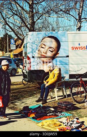 Winter flea market in sunlight, a man selling old clothes beside a van with a large ad showing a model’s face splashing water; analog cross-processed Stock Photo