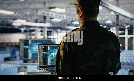 Army programmer in high tech server hub uses AI application on laptop to review reconnaissance information. Data center IT expert wearing military uniform uses AI on notebook to plan mission, camera B Stock Photo