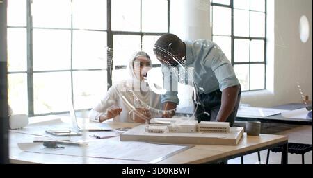 Collaborating Indian woman wearing hijab, colleague examining scale model with blueprints at studio Stock Photo