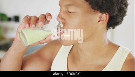 Drinking man wearing white tank top holding glass with green smoothie in home kitchen, with plant Stock Photo