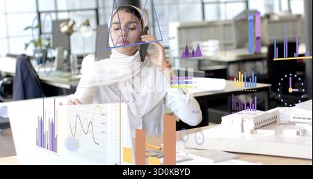 Architect wearing blouse talking on phone and analyzing charts on laptop in office with scale model Stock Photo