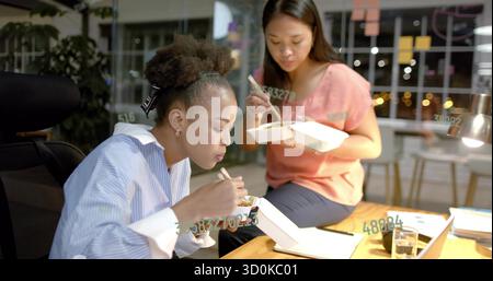 Eating lunch two colleagues using chopsticks at desk in office workspace, with takeout and laptop Stock Photo