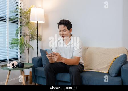 A young man sitting on a sofa using his smartphone at home. He looks relaxed and focused while interacting online, possibly chatting, reading, or mana Stock Photo