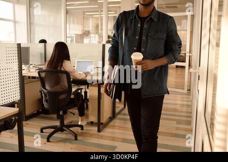 End of working day. Unrecognizable black man leaving office with coffee to go, unrecognizable woman working on laptop behind Stock Photo